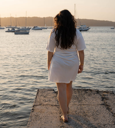 A woman with long dark curly hair stands on a concrete dock by the water, viewed from behind, wearing a short white dress. Sailboats are anchored in the calm water under a warm golden sunset sky.