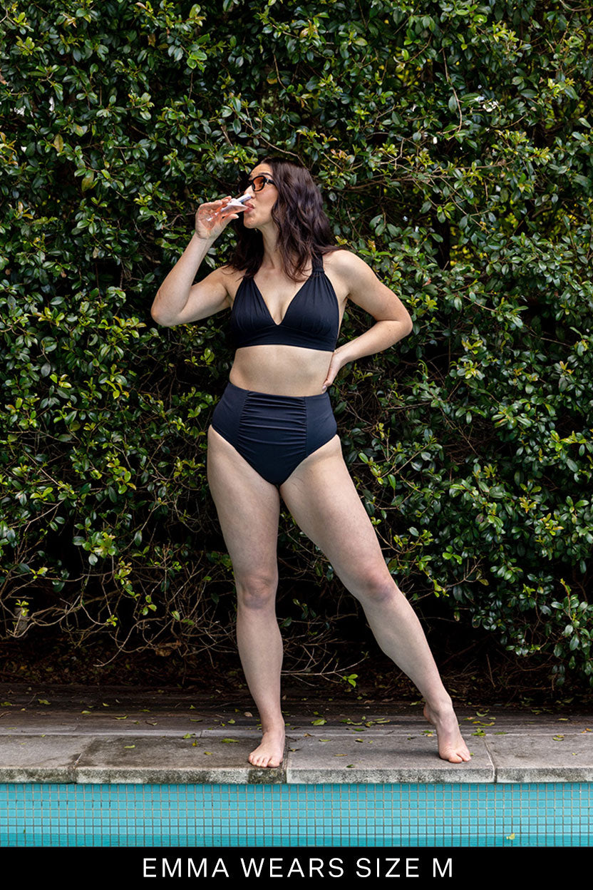 A woman stands by a pool in Fearless au’s All Day Bikini Bottom, sipping from a can and wearing sunglasses. She poses confidently before lush greenery. Text at the bottom reads, “EMMA WEARS SIZE M.”.