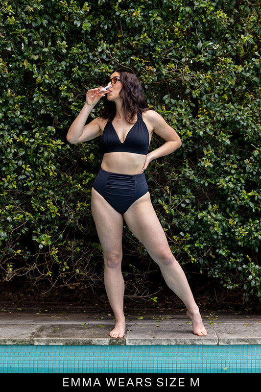 A woman stands by a pool in Fearless au’s All Day Bikini Bottom, sipping from a can and wearing sunglasses. She poses confidently before lush greenery. Text at the bottom reads, “EMMA WEARS SIZE M.”.