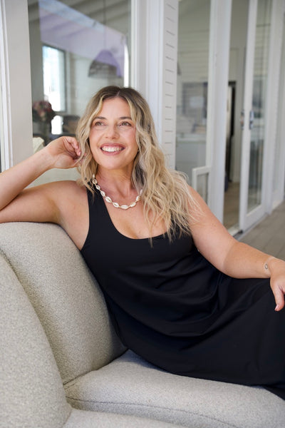 A smiling blonde woman wearing a black sleeveless dress and a white shell necklace, leaning back on a light beige sofa in a bright, modern room.