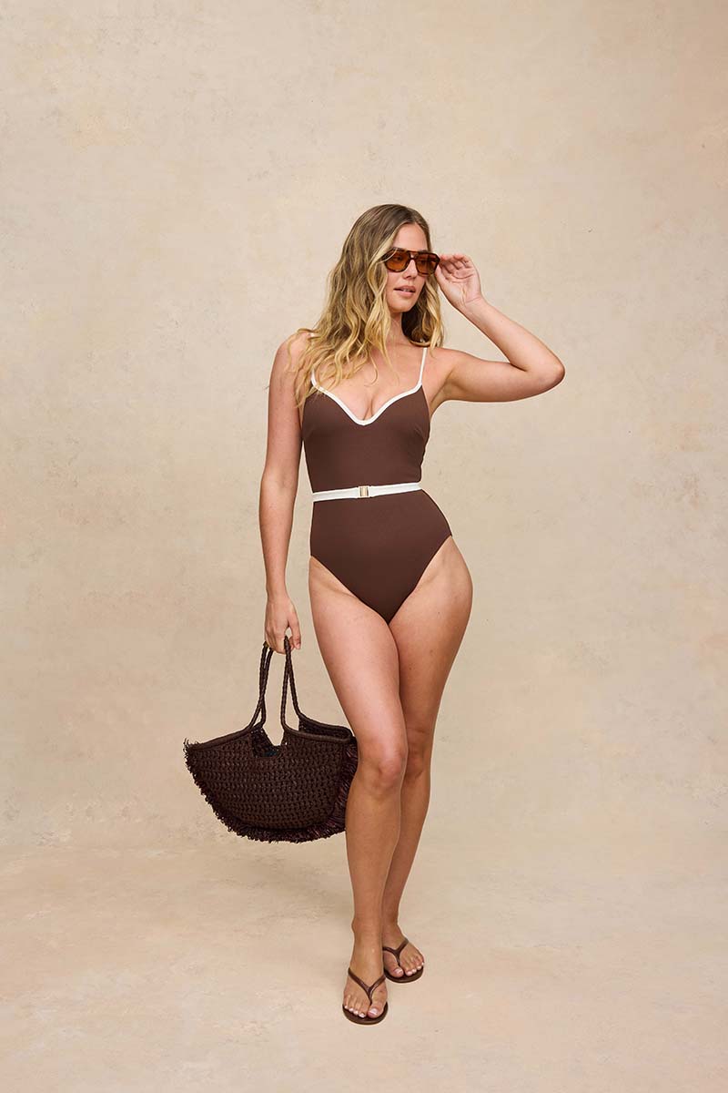 A woman modeling a brown one-piece swimsuit with white trim and a thin white waist belt, standing against a textured beige studio backdrop and holding a dark woven tote bag.
