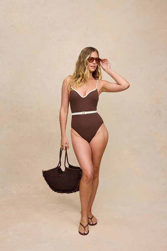 A woman modeling a brown one-piece swimsuit with white trim and a thin white waist belt, standing against a textured beige studio backdrop and holding a dark woven tote bag.
