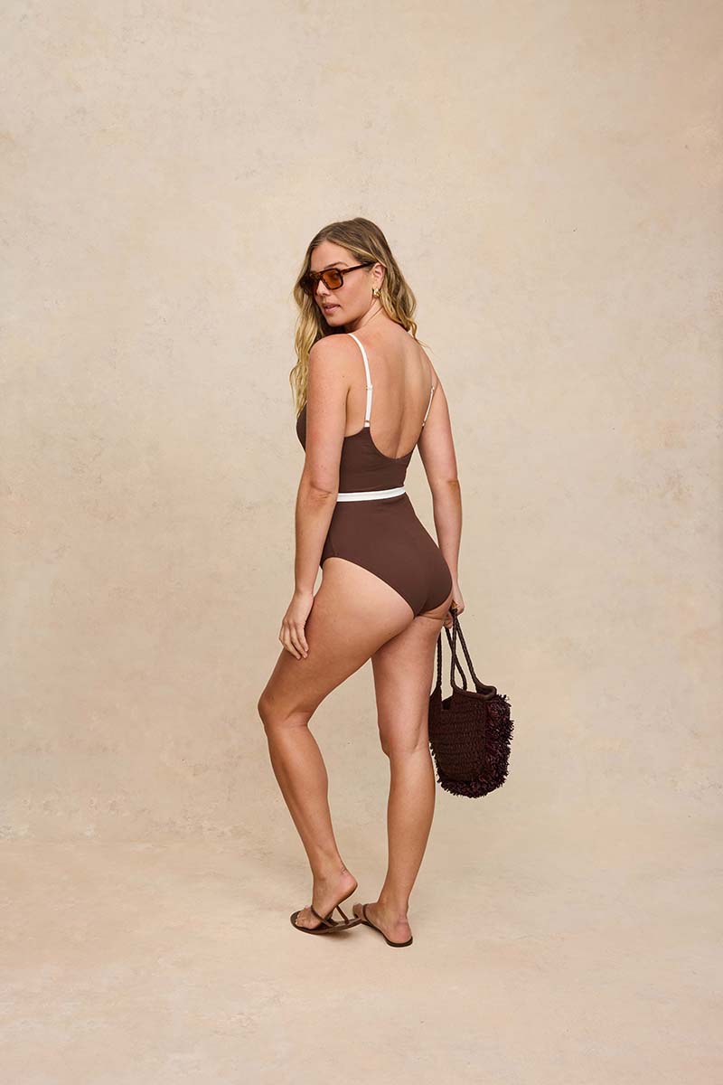 A woman modeling a brown one-piece swimsuit with thin white shoulder straps and a white waistband, standing against a textured beige studio backdrop and holding a dark brown fuzzy handbag.