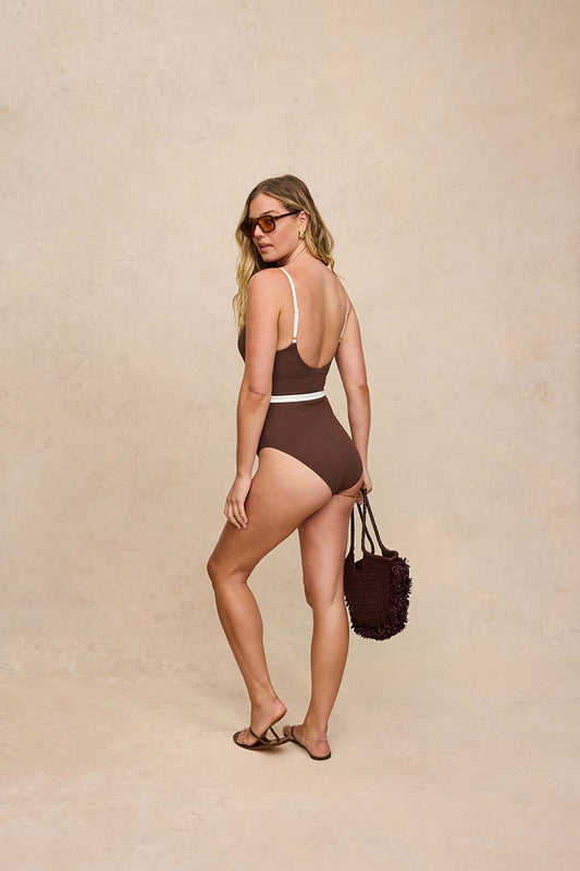 A woman modeling a brown one-piece swimsuit with thin white shoulder straps and a white waistband, standing against a textured beige studio backdrop and holding a dark brown fuzzy handbag.
