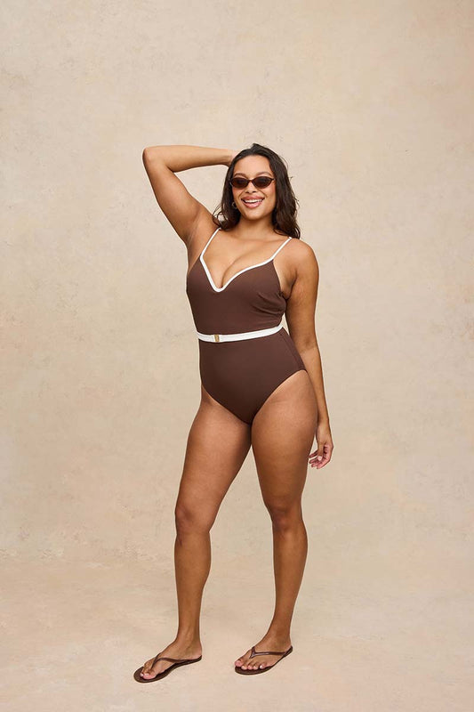 A woman modeling a brown one-piece swimsuit with white contrast trim and a matching belt, standing against a textured beige studio backdrop.