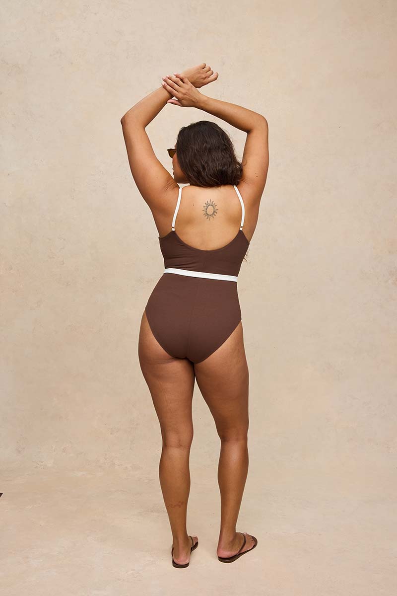 A woman viewed from behind wearing a one-piece swimsuit in dark brown with thin white shoulder straps and a white waistband, standing against a textured beige wall.