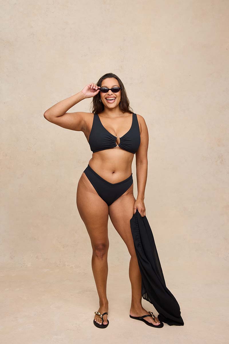 A woman modeling a black two-piece bikini with a front keyhole cutout, wearing black sunglasses and holding a matching black swim cover-up against a textured beige studio backdrop.