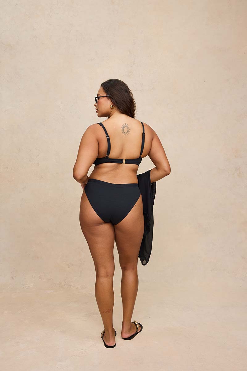 A woman stands with her back to the camera wearing a black string bikini and holding a black cover-up against a textured beige studio backdrop.