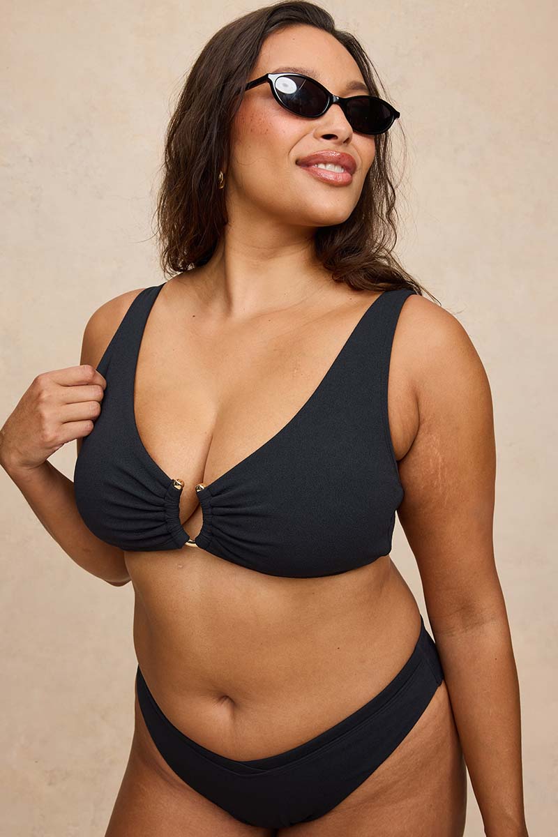 A woman modeling a black two-piece bikini with a keyhole front and matching black sunglasses, posed against a neutral beige textured background.