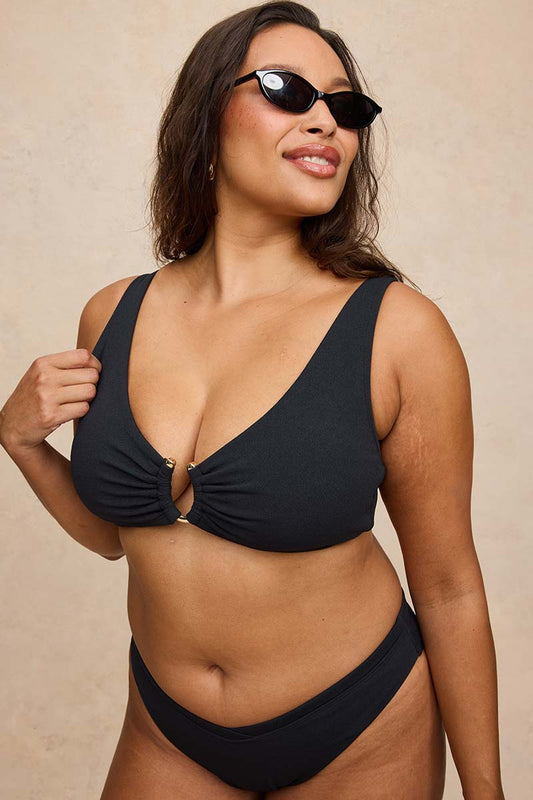 A woman modeling a black two-piece bikini with a keyhole front and matching black sunglasses, posed against a neutral beige textured background.