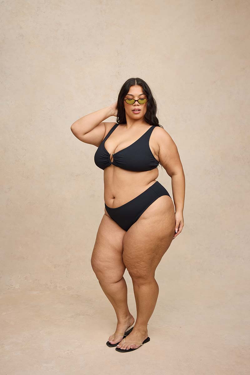 A plus-size woman modeling a black two-piece bikini with a front keyhole detail, standing against a textured beige studio backdrop.