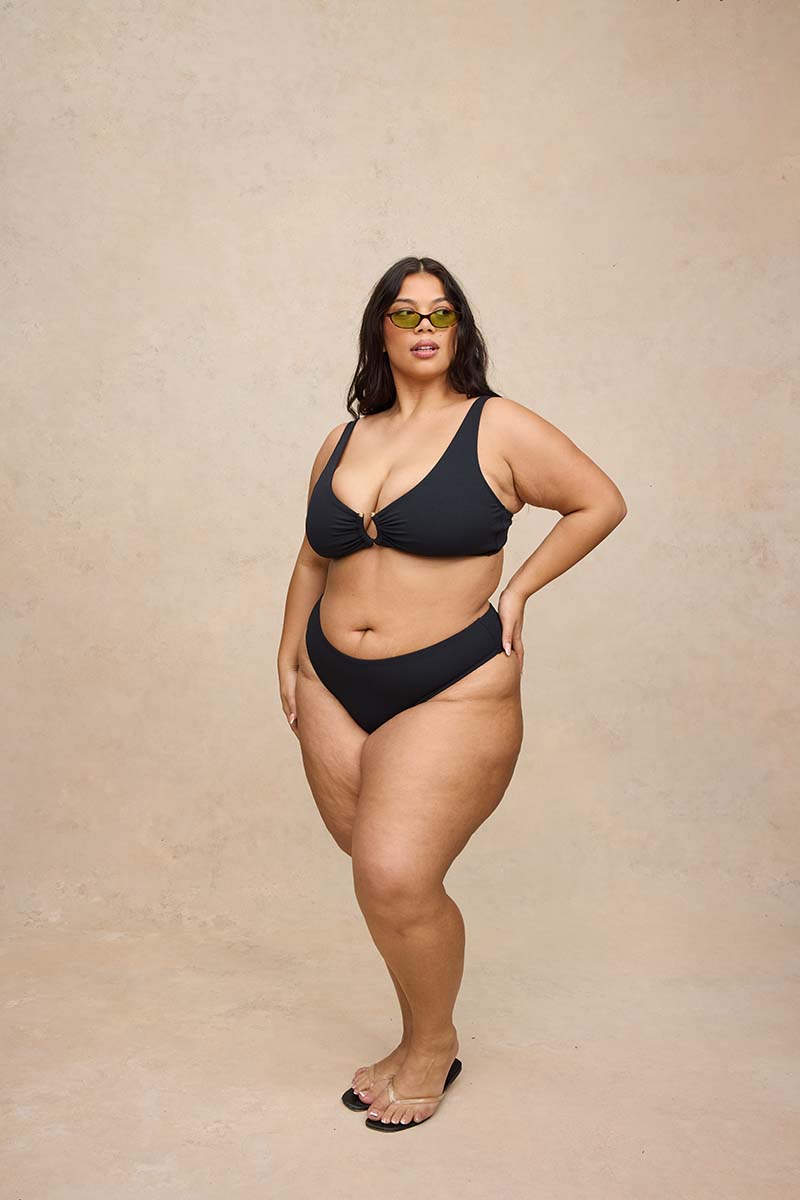 A woman modeling a black two-piece bikini with a front keyhole detail on the top, paired with yellow-tinted sunglasses and black sandals, standing against a textured beige studio backdrop.