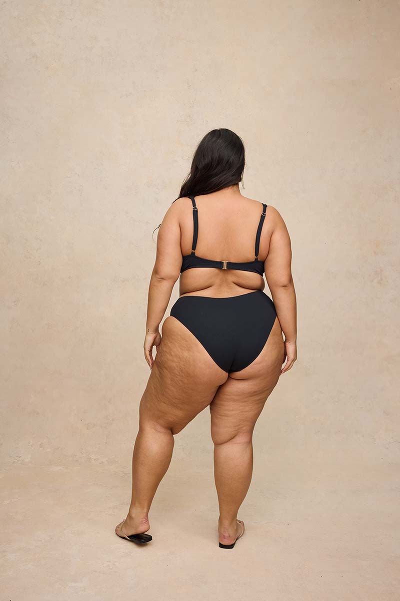 A woman is shown from behind wearing a black two-piece bikini with a small gold clasp at the center back, standing against a textured beige studio backdrop.