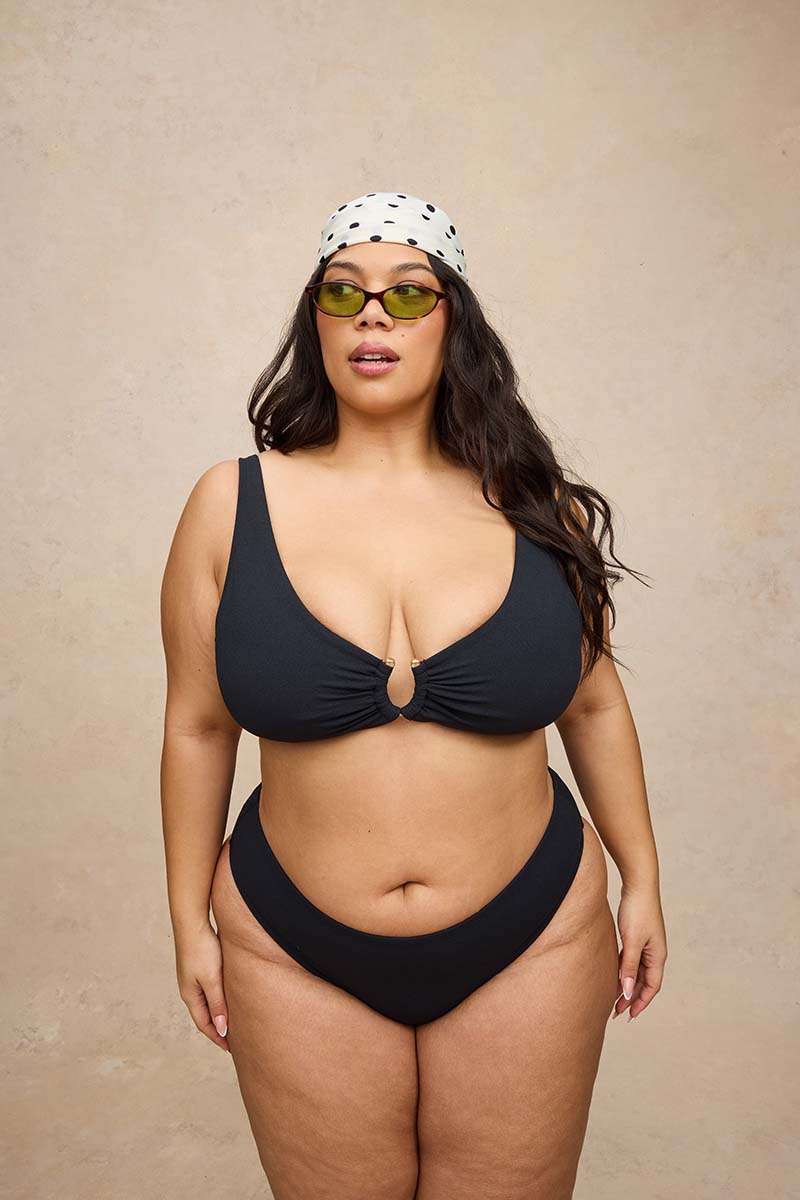 A woman modeling a black two-piece bikini with a front keyhole cutout, styled with a white bandana and green-tinted sunglasses against a textured beige background.