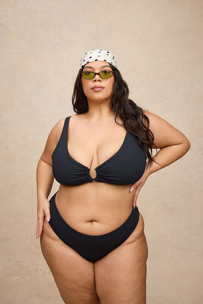 A woman modeling a black two-piece bikini with a keyhole cutout at the center of the top, paired with a white bandana and green-tinted sunglasses, against a textured beige background.