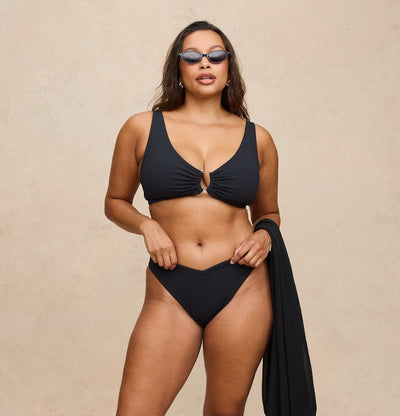 A woman modeling a black two-piece bikini with a front keyhole cutout on the top, paired with matching high-cut bottoms and a draped black swim cover-up, set against a textured beige studio backdrop.