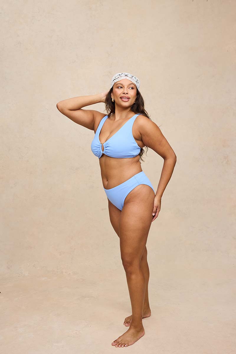 A woman modeling a light blue two-piece bikini with a halter top and high-waisted bottoms, paired with a white patterned headband, standing against a textured beige studio backdrop.
