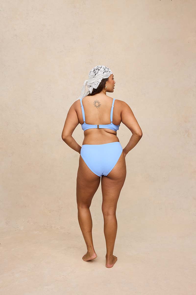 A woman stands with her back to the camera wearing a light blue two-piece bikini and a white patterned headscarf, set against a textured beige studio backdrop.
