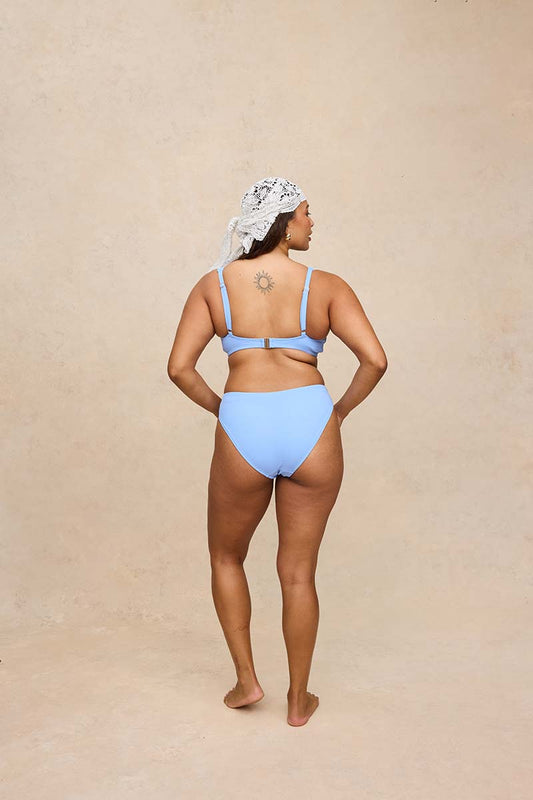 A woman stands with her back to the camera wearing a light blue two-piece bikini and a white patterned headscarf, set against a textured beige studio backdrop.