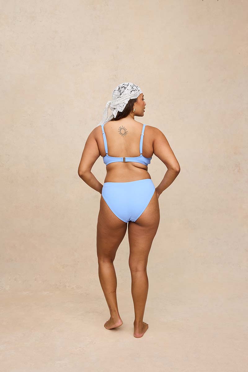 A woman stands with her back to the camera wearing a light blue two-piece bikini and a white patterned headscarf, set against a textured beige studio backdrop.
