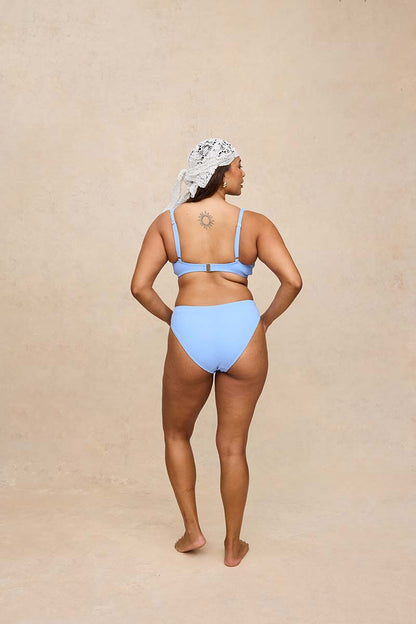 A woman stands with her back to the camera wearing a light blue two-piece bikini and a white patterned headscarf, set against a textured beige studio backdrop.