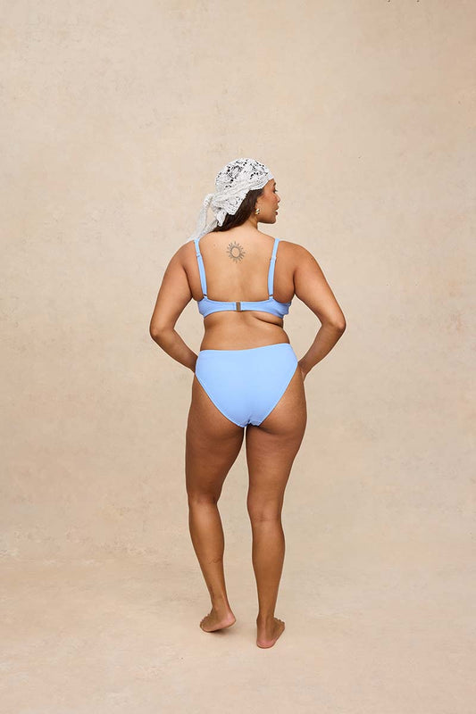 A woman stands with her back to the camera wearing a light blue two-piece bikini and a white patterned headscarf, set against a textured beige studio backdrop.