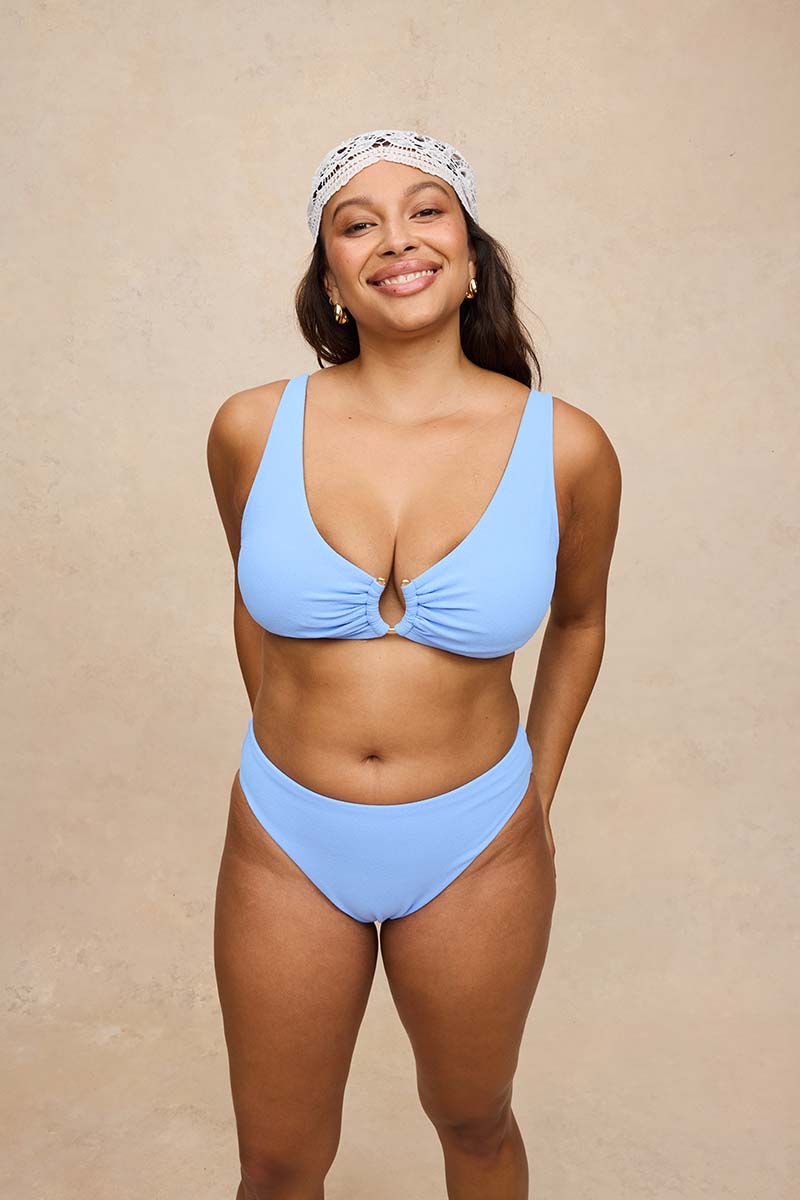 A woman modeling a light blue two-piece bikini with a keyhole front on the top, paired with a white patterned headband, against a textured beige studio backdrop.