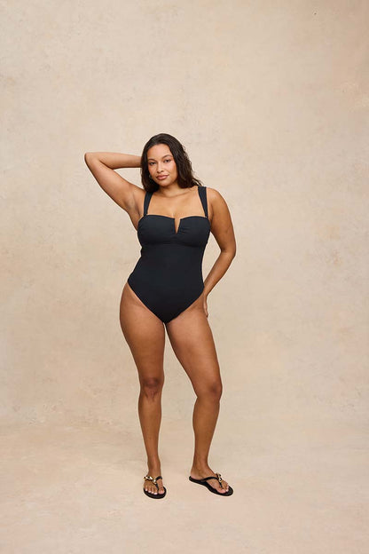 A woman modeling a black one-piece swimsuit with a sweetheart neckline and keyhole cutout, standing against a textured beige studio backdrop.