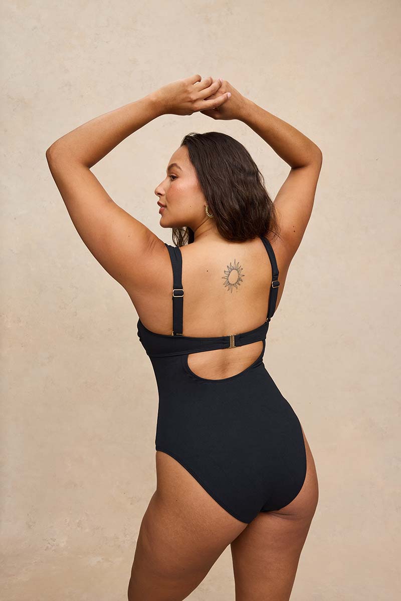 A woman modeling a black one-piece swimsuit from the back, showing the cutout and adjustable straps, against a textured beige background.