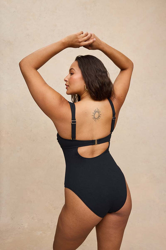 A woman modeling a black one-piece swimsuit from the back, showing the cutout and adjustable straps, against a textured beige background.