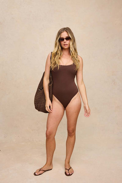 A woman modeling a ribbed brown one-piece swimsuit, styled with a large woven brown tote bag and dark sunglasses, standing against a textured beige studio backdrop.