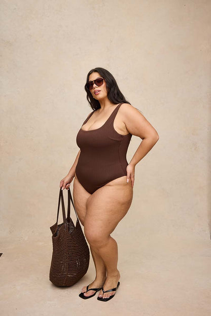 A plus-size woman modeling a ribbed brown one-piece swimsuit, holding a brown woven tote bag and wearing black flip-flops against a textured beige studio backdrop.