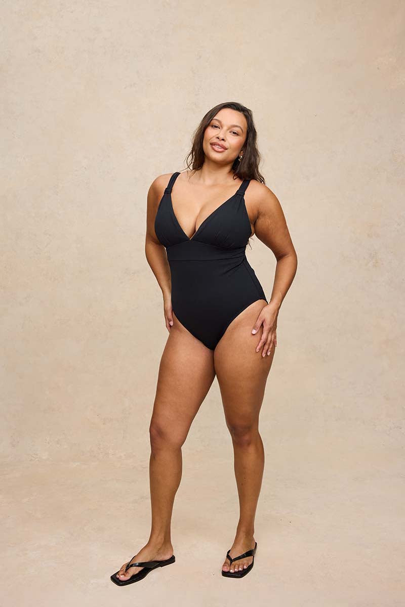 A woman modeling a black one-piece swimsuit with a deep V-neckline, standing against a textured beige studio backdrop.