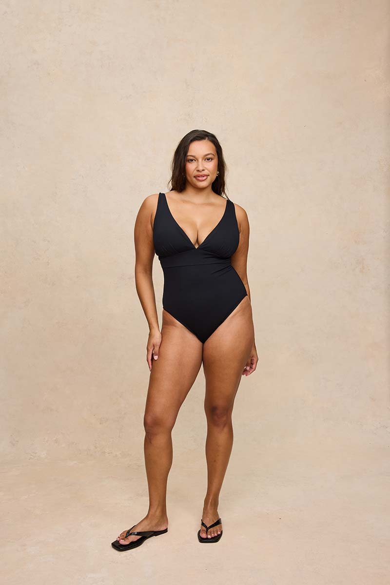 A woman modeling a black one-piece swimsuit with a deep V-neckline and shoulder straps, standing against a textured beige studio backdrop.
