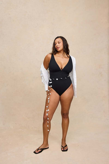 A woman modeling a black one-piece swimsuit with silver shell embellishments down the side, styled with an open white shirt and black flip-flops against a beige textured studio backdrop.