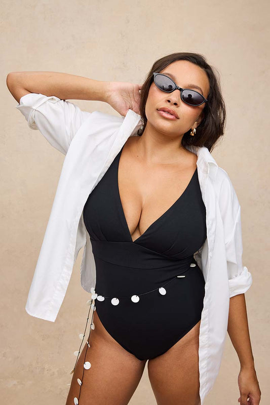 A woman modeling a black one-piece swimsuit with a wrap-style V-neckline, styled with an open white button-down shirt and dark sunglasses against a neutral beige background.