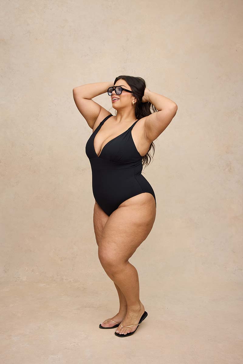 A plus-size woman modeling a black one-piece swimsuit with a deep V-neckline and sunglasses, standing against a textured beige studio backdrop.
