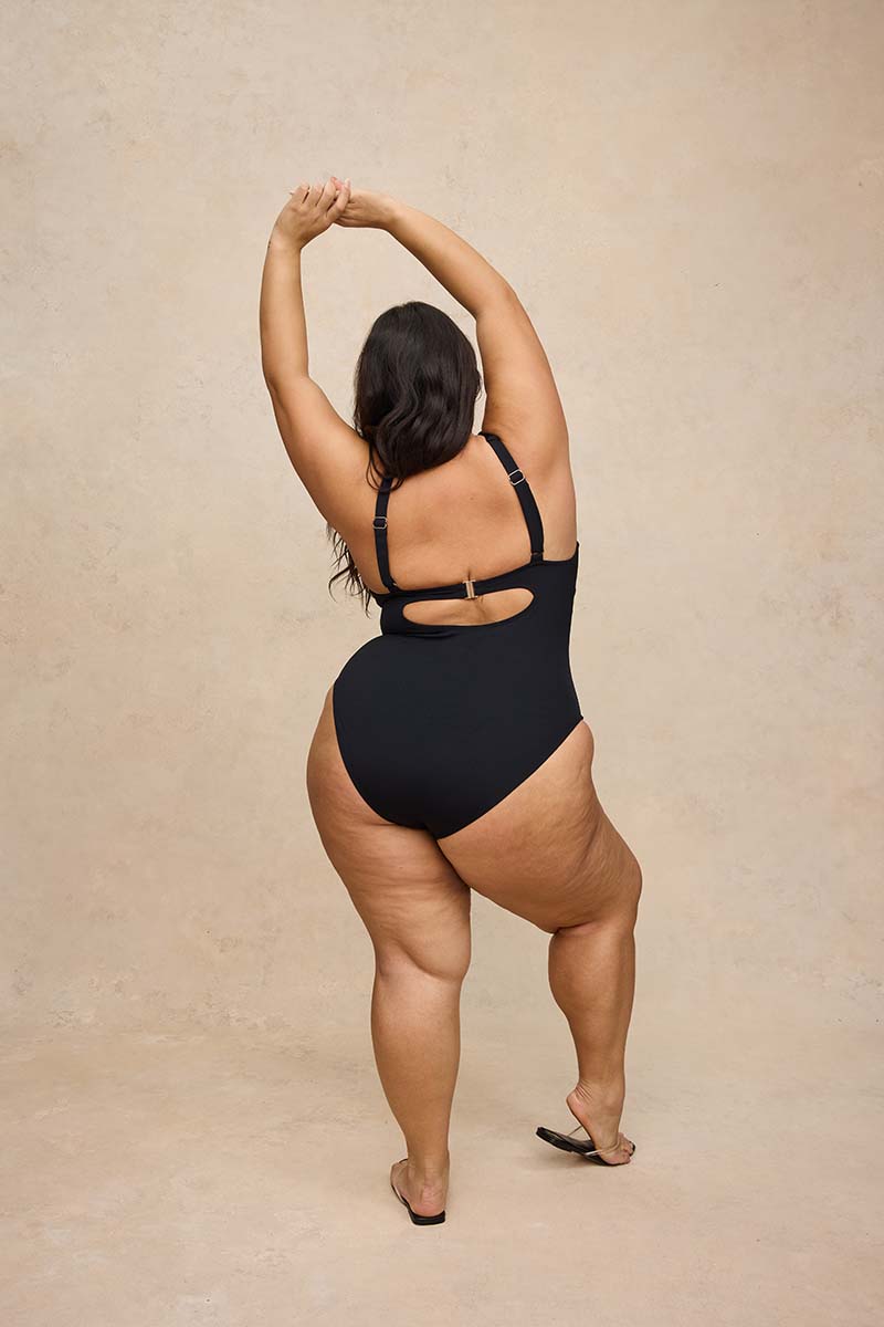 A woman wearing a black one-piece swimsuit is photographed from behind against a warm beige textured studio backdrop. She has her arms raised overhead and is wearing black sandals.