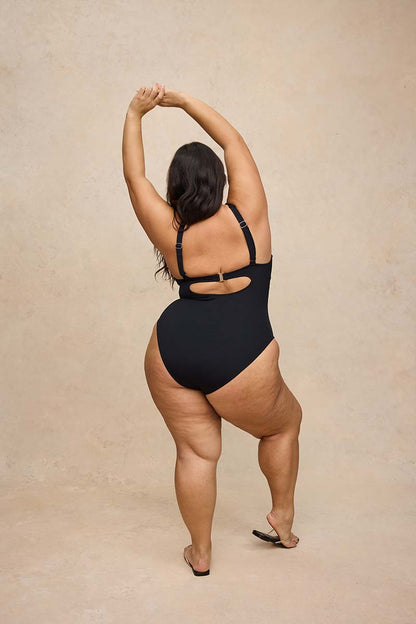 A woman wearing a black one-piece swimsuit is photographed from behind against a warm beige textured studio backdrop. She has her arms raised overhead and is wearing black sandals.