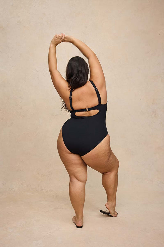 A woman wearing a black one-piece swimsuit is photographed from behind against a warm beige textured studio backdrop. She has her arms raised overhead and is wearing black sandals.