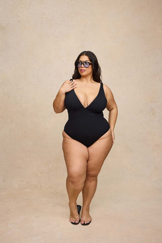 A plus-size model wearing a black one-piece swimsuit and black-framed sunglasses, standing against a textured beige studio backdrop.