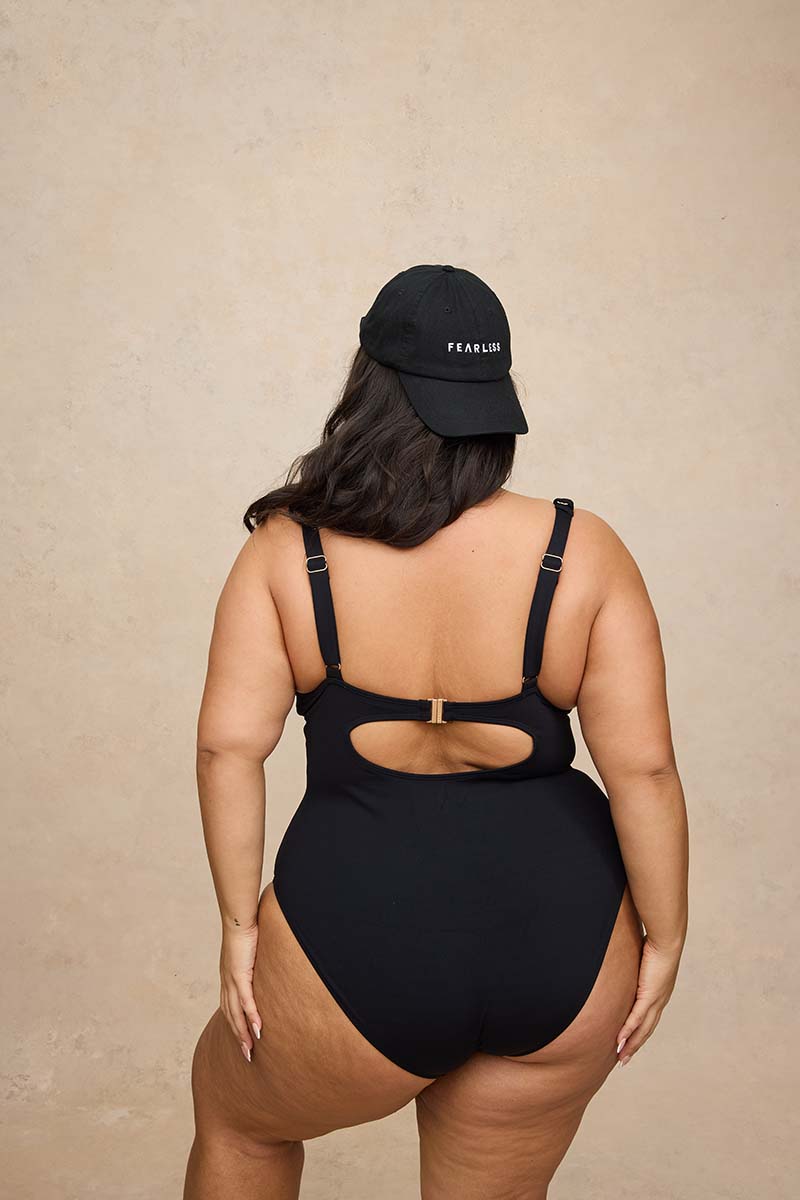 A woman shown from the back wearing a black one-piece swimsuit with a cutout at the back and a black baseball cap with white text, standing against a textured beige wall.
