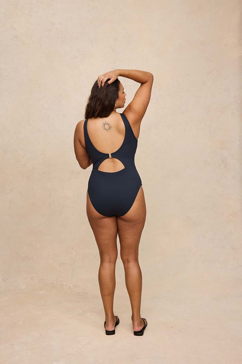 A woman stands with her back to the camera wearing a black one-piece swimsuit with a cutout at the center of the back, set against a textured beige studio backdrop.