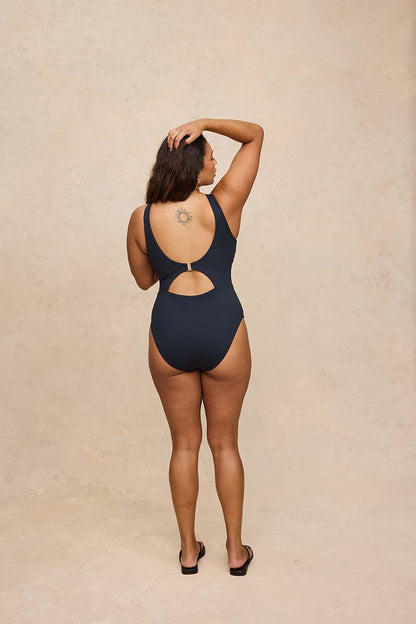 A woman stands with her back to the camera wearing a black one-piece swimsuit with a cutout at the center of the back, set against a textured beige studio backdrop.