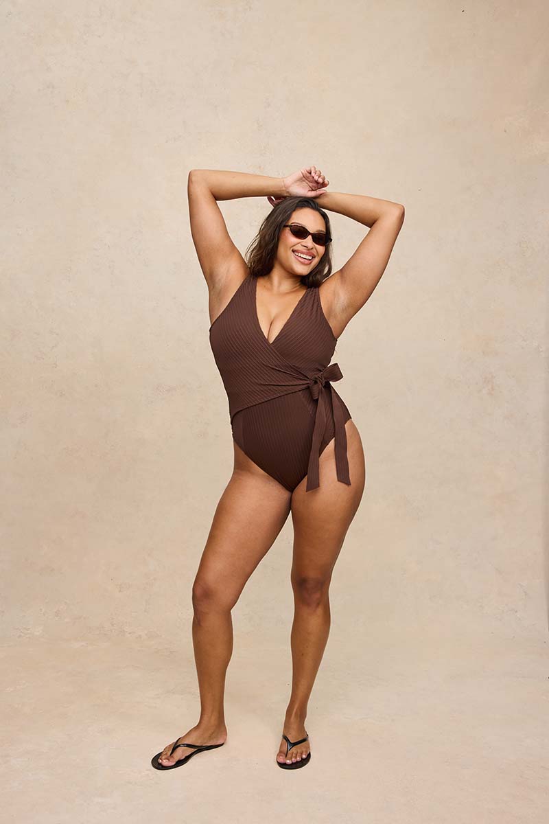 A woman modeling a ribbed brown one-piece swimsuit with a wrap-front tie detail, standing against a textured beige studio backdrop.