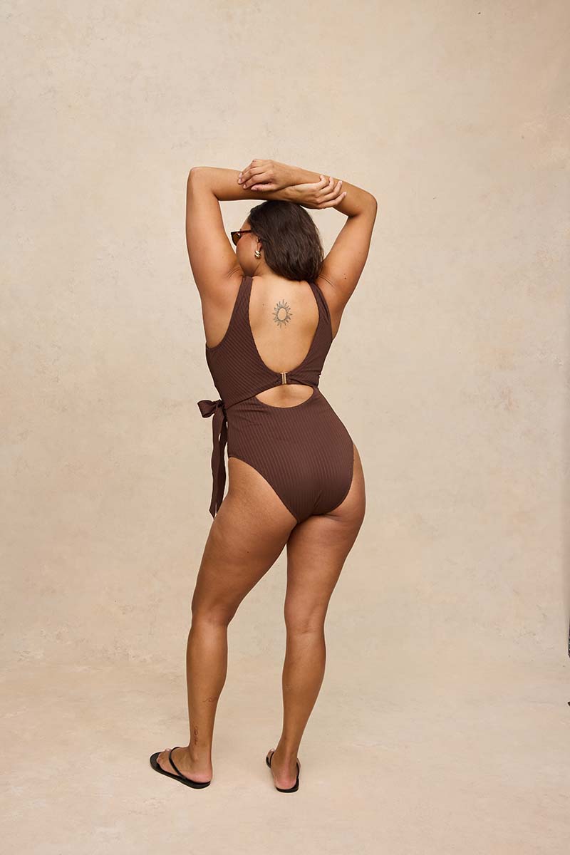 A woman wearing a dark brown one-piece swimsuit, viewed from behind, posing with her arms raised against a textured beige studio backdrop.