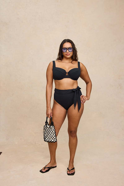 A woman modeling a black two-piece bikini with a bralette top and high-waisted side-tie bottoms, standing against a textured beige studio backdrop and holding a black-and-white patterned handbag.