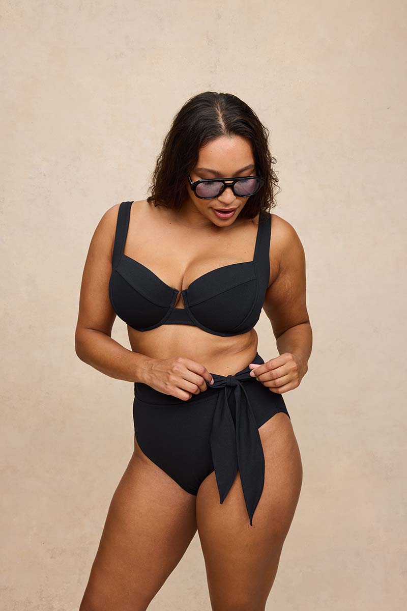 A woman modeling a black two-piece swimsuit with a bra-style top and high-waisted tie-front bottoms, standing against a textured beige studio backdrop.