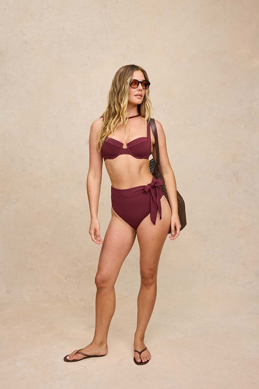 A woman modeling a two-piece bikini in a deep burgundy/maroon color, standing against a textured beige studio backdrop. She is wearing sunglasses, a brown shoulder bag, and dark flip-flop sandals.
