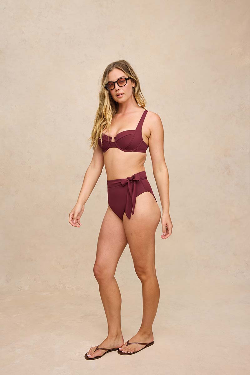 A woman modeling a two-piece high-waisted bikini in a deep burgundy color, paired with dark sunglasses and flip-flops, standing against a textured beige studio backdrop.